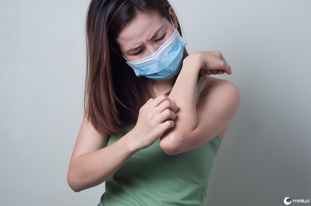 young asian woman scratching arm while wearing surgical mask
